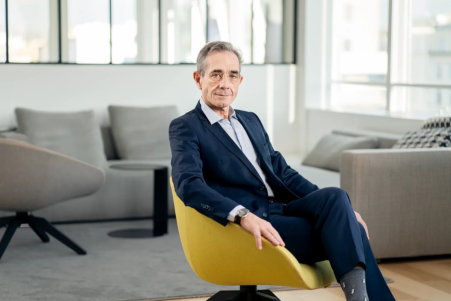 Portrait d’Un Dirigeant d’Entreprise à Paris Ce portrait corporate présente un dirigeant d'entreprise assis élégamment dans un bureau moderne et lumineux à Paris. Il porte un costume bleu classique et un sourire confiant, dégageant une image professionnelle. Ce portrait à été réalisé par un photographe spécialisé dans le portrait d'entreprise à Paris.